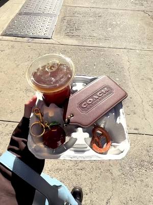 A post by @coach on TikTok caption: We’ll take one coffee, an XL Hangtag, a Small Apple Bag Charm, and Pretzel Bag Charm to-go 🥨🍎 #CoachNY