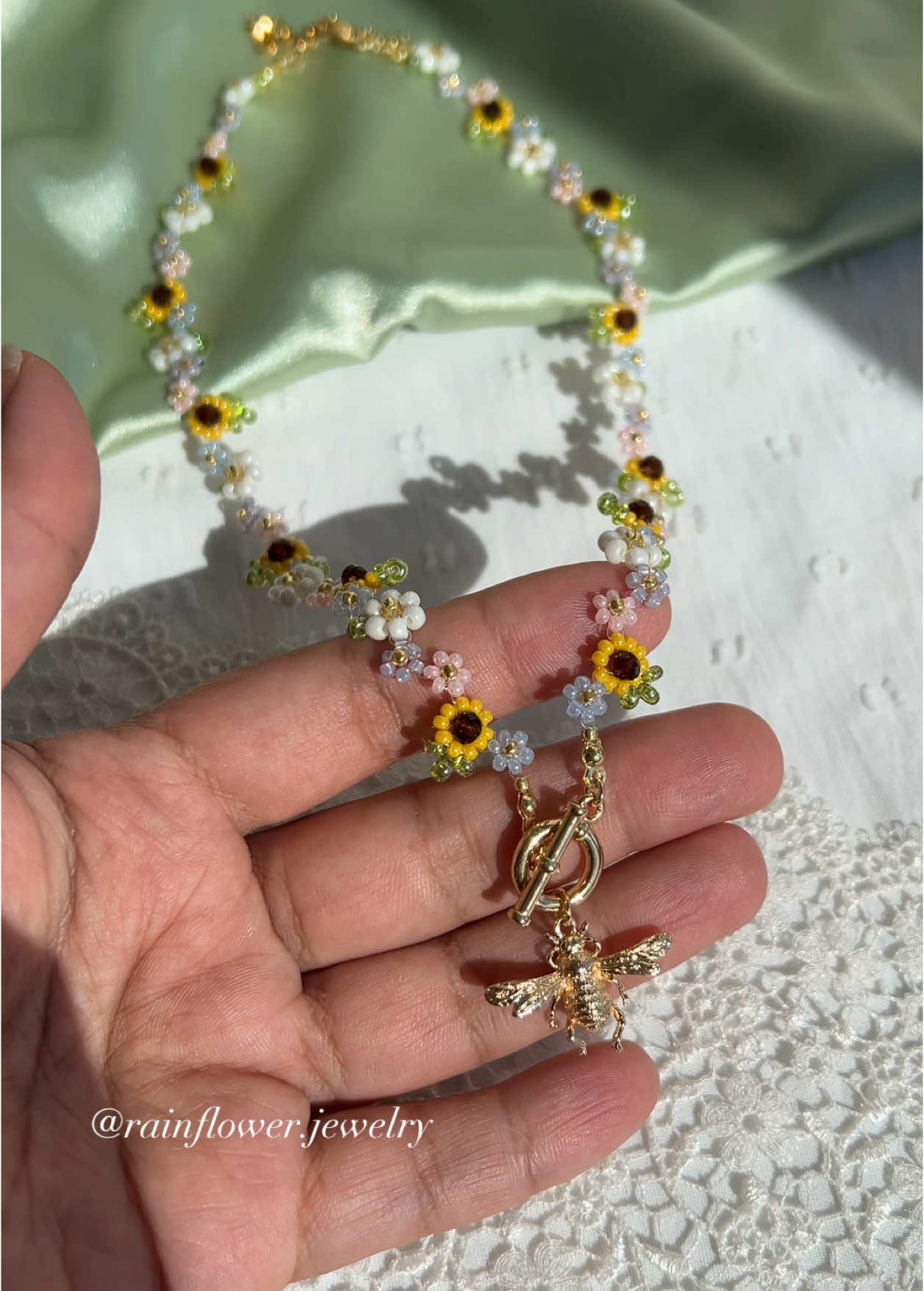 A post by @rainflower.jewelry on TikTok caption: I finally made a sunflower necklace with a tiny bee charm — and I’m obsessed! It looks like pure sunshine and feels like summer🥹🌻🌱✨ Now available in my shop with free worldwide shipping! (Link in bio)💞 #sunflowernecklace #beejewelry #handmadejewelry #summervibes #aestheticnecklace #cottagecorejewelry #floralaccessories #daintyjewelry #smallbiztok #shopsmall #supporthandmade #handmadewithlove #trendingjewelry 